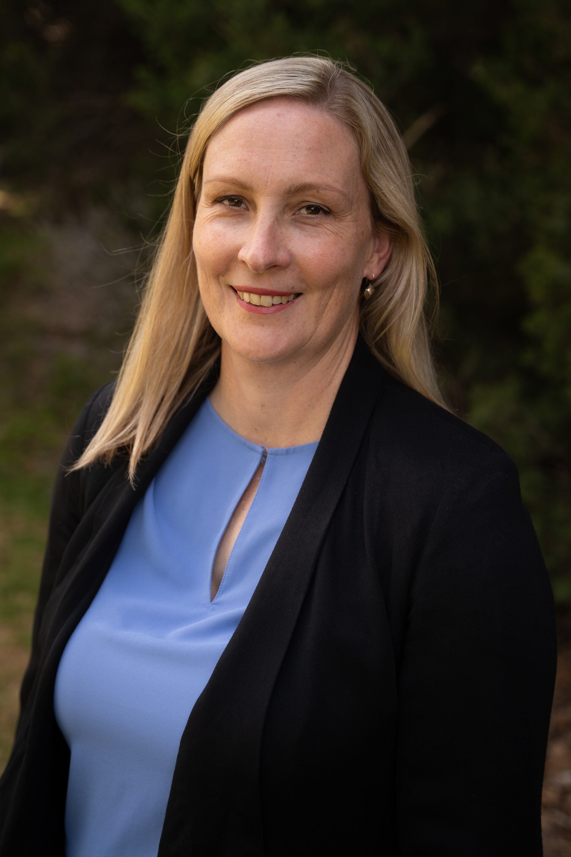 Woman with long blonde hair in a blue top and black blazer, smiling outdoors with greenery behind her.