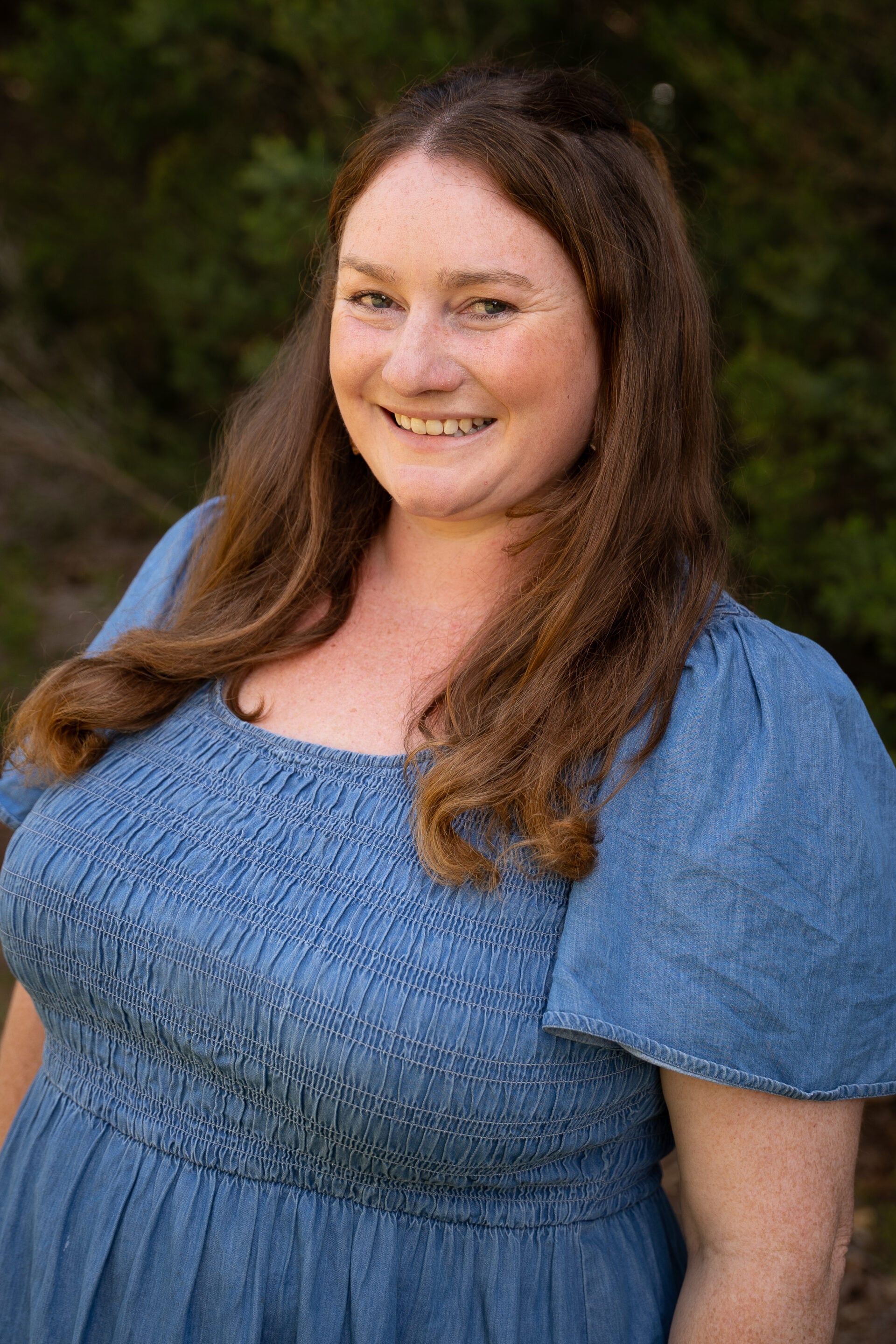 Woman with long brown hair wearing a blue dress, smiling outdoors with greenery in the background.