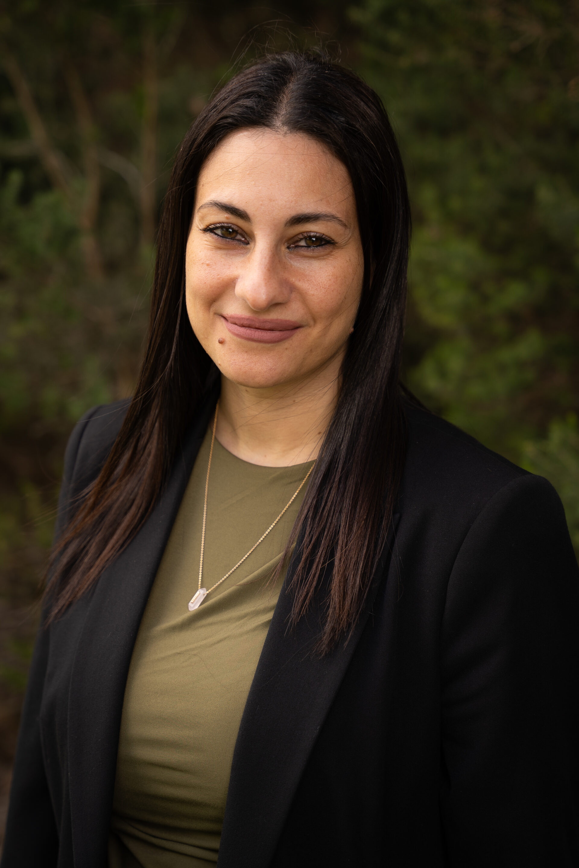 Woman with long dark hair in a black blazer and green top smiling outdoors with greenery in the background.