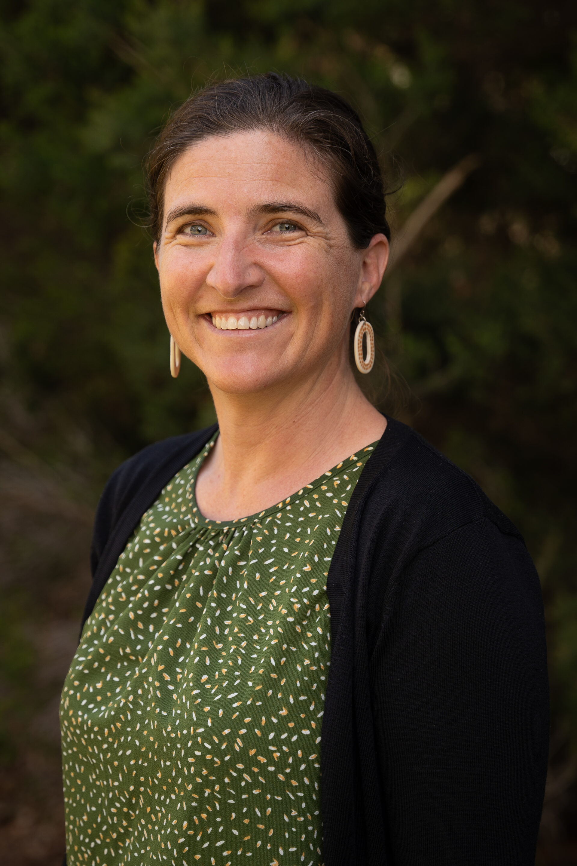 Woman with brown hair in a bun, wearing a green patterned top and black cardigan, smiling outdoors.