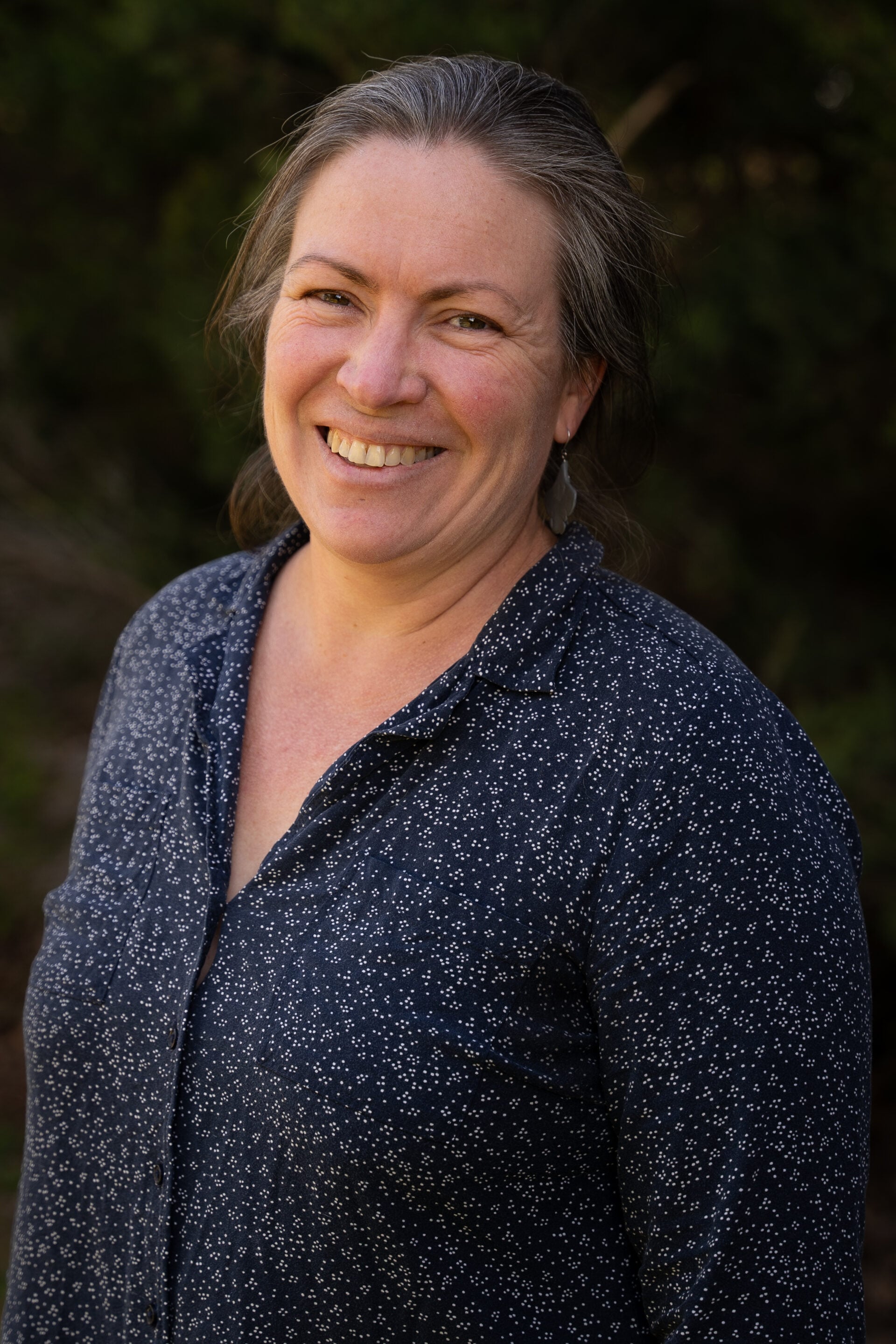 Woman with grey hair smiling outdoors, wearing a navy blue shirt with a white spotted pattern.