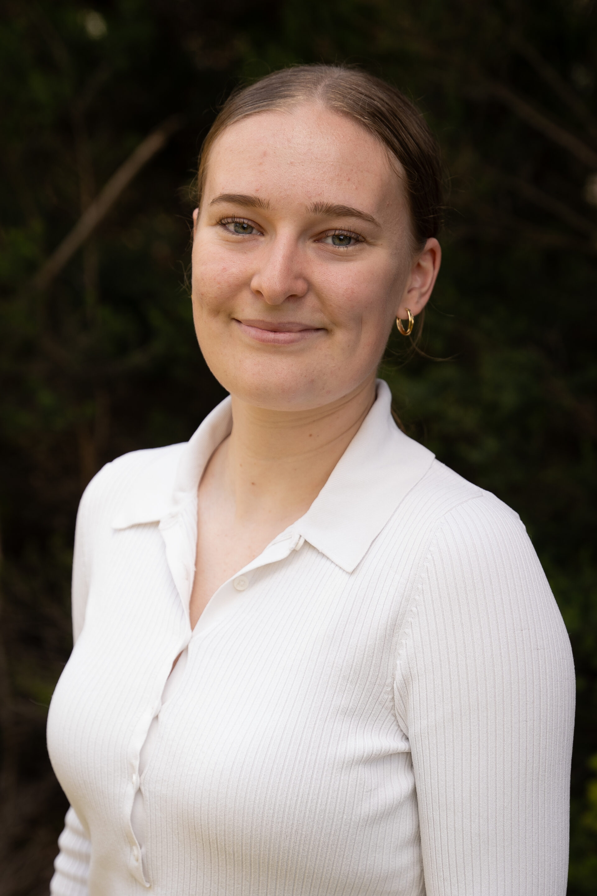 Woman in a white shirt standing outdoors, with greenery in the background.