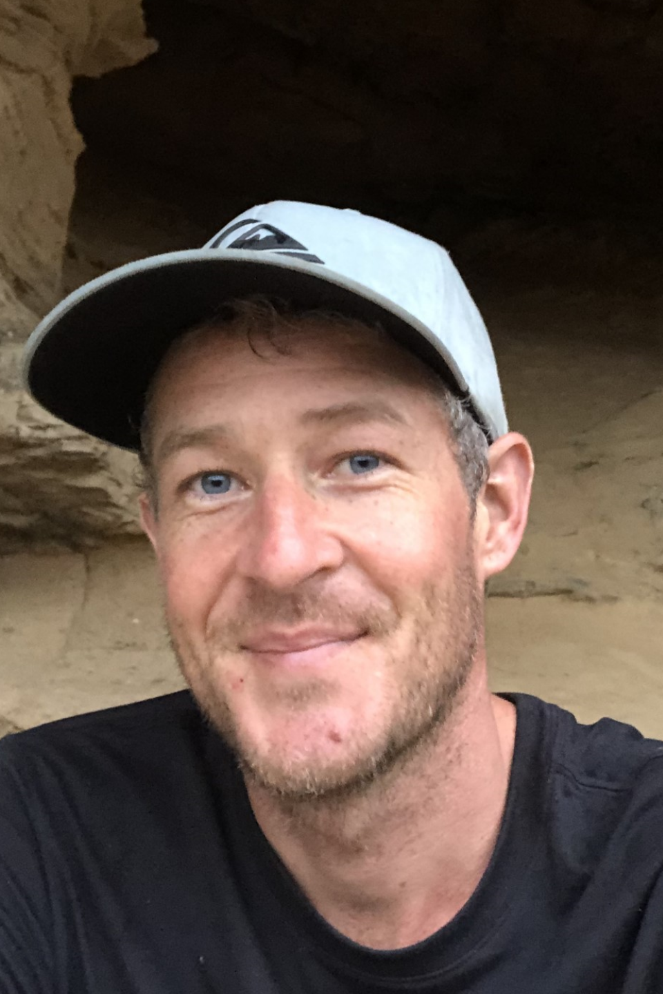 Man with blue eyes wearing a grey cap, smiling in front of a rocky background.