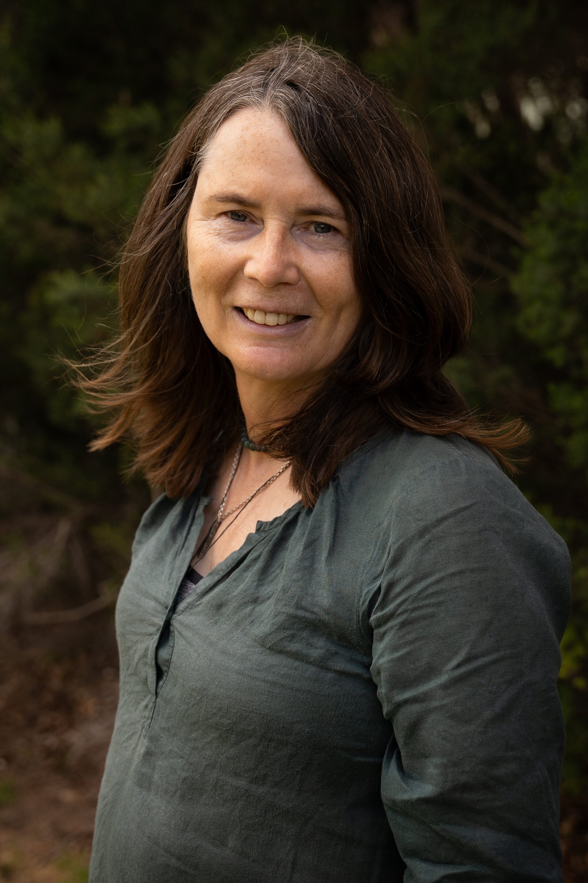 Woman with long brown hair in a green shirt, smiling outdoors with greenery in the background.