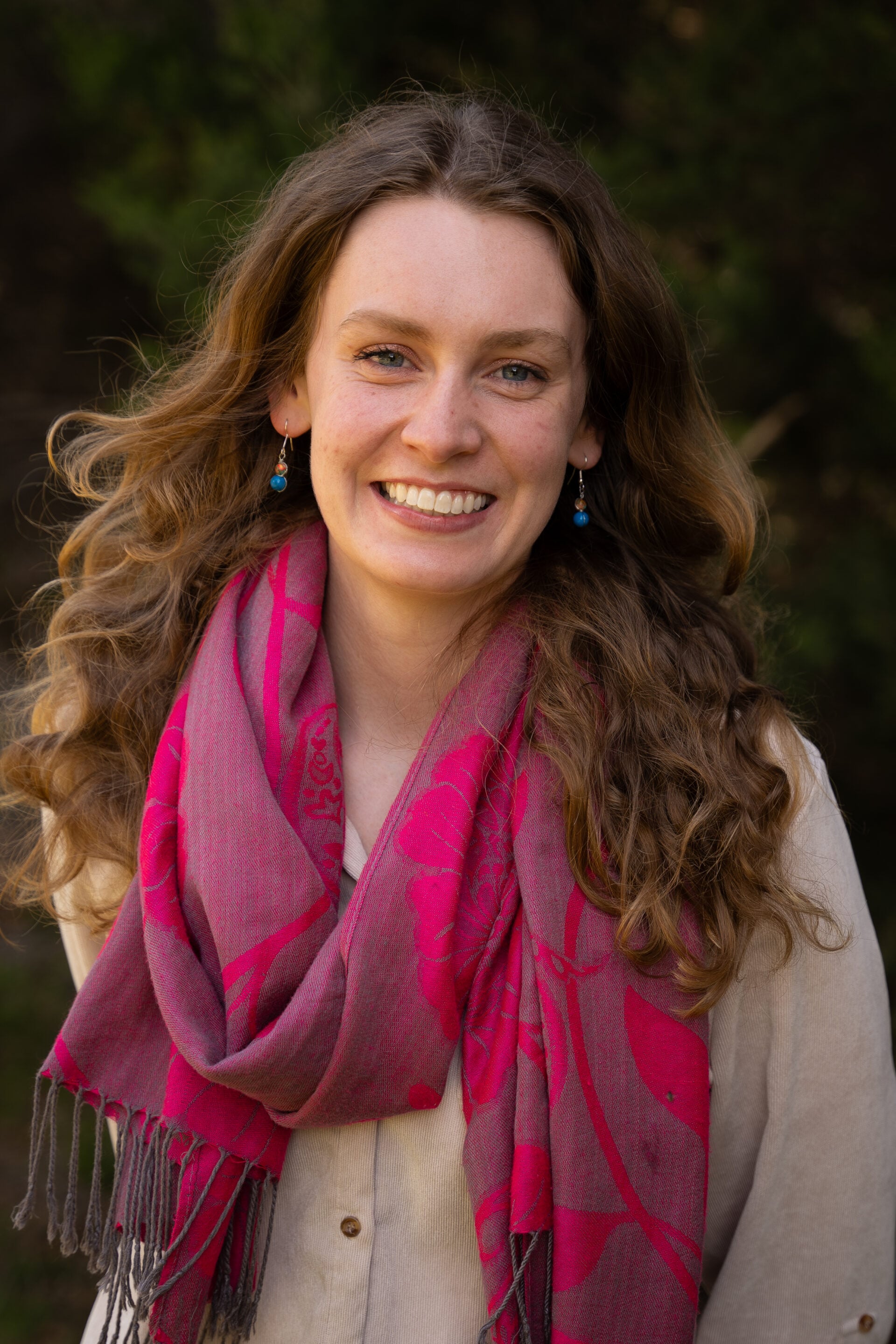 A woman with wavy hair wears a pink scarf and earrings, standing outdoors with greenery in the background.