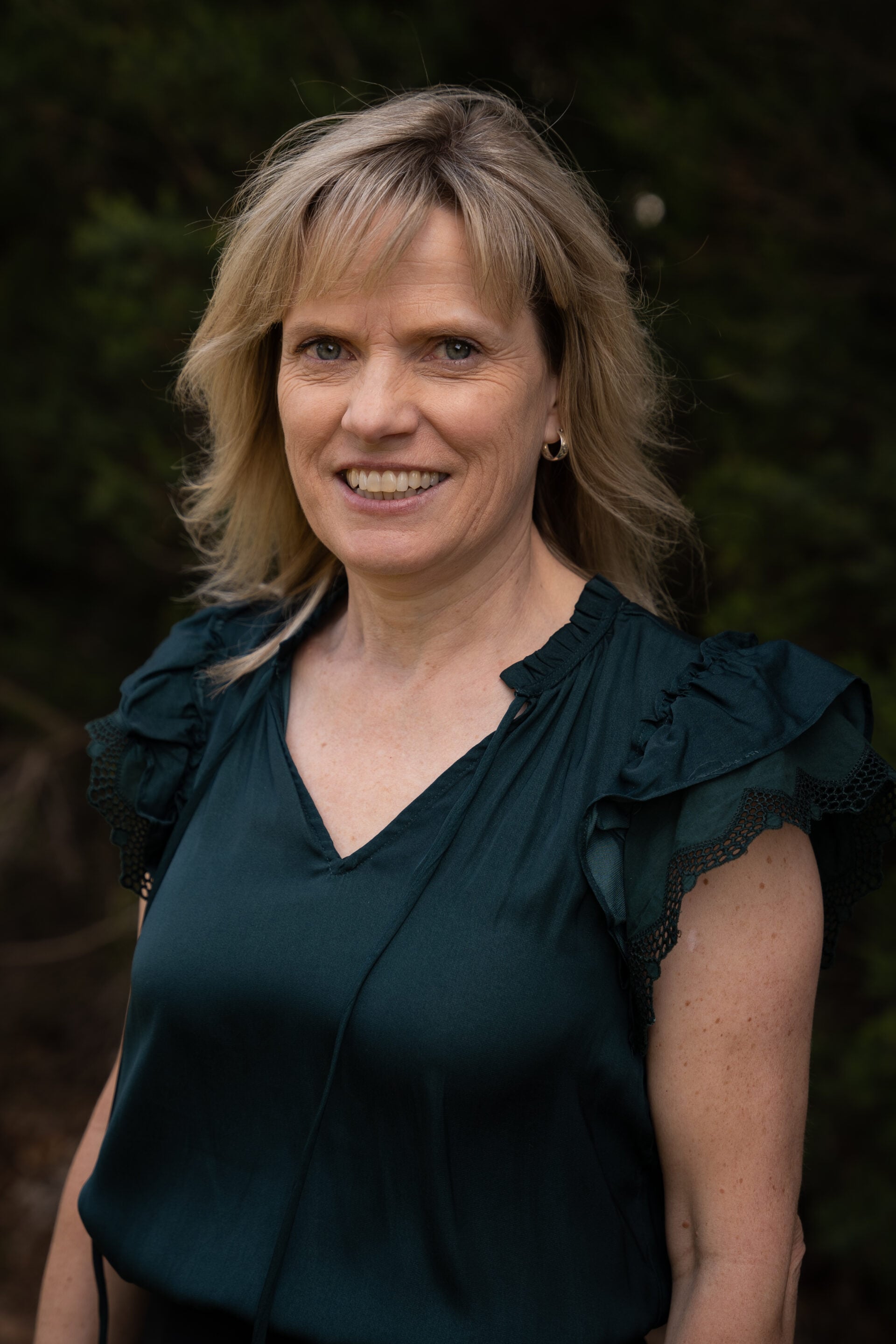 Woman with blonde hair wearing a green blouse, smiling outdoors in front of dark greenery.