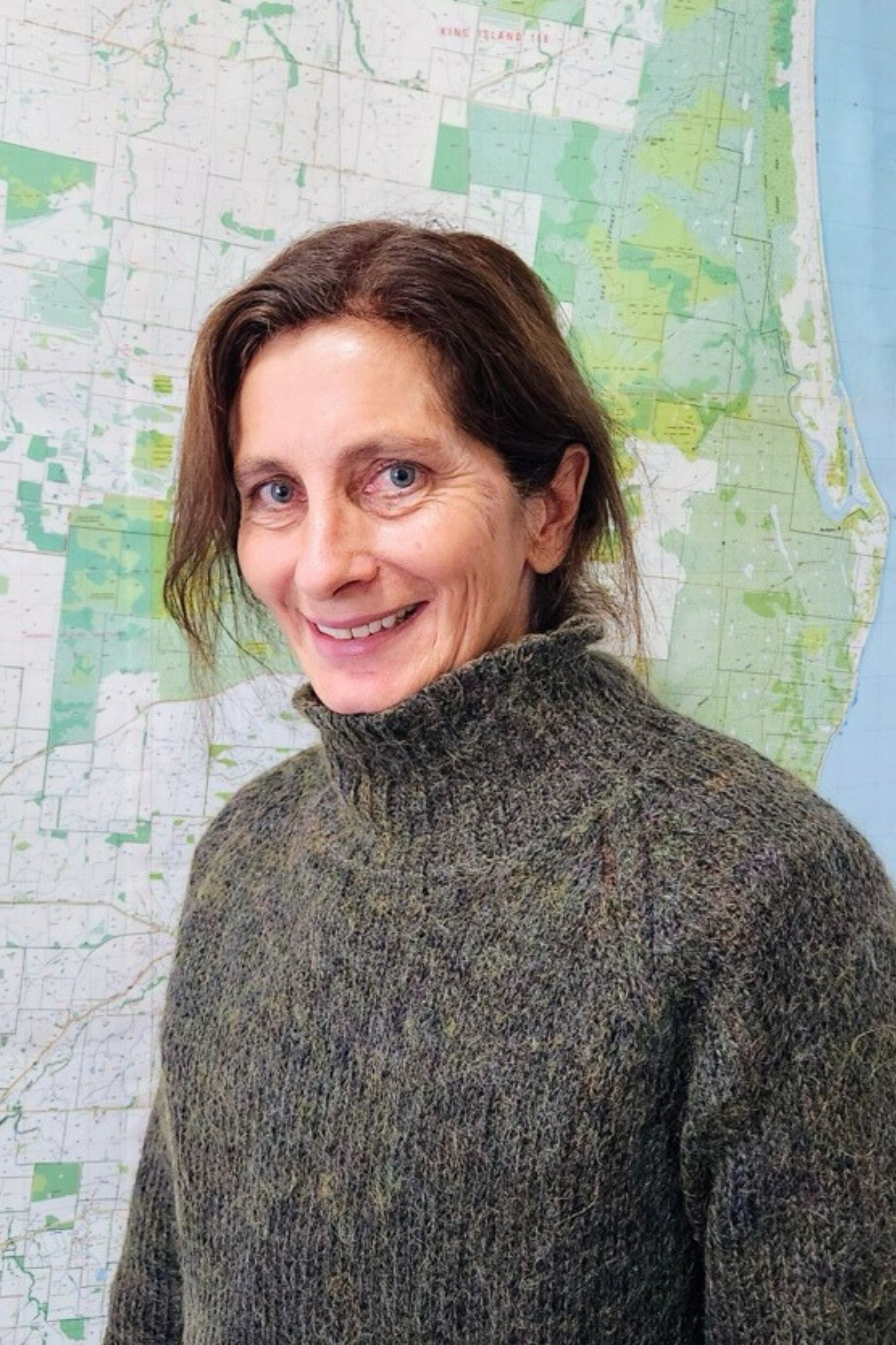 Woman with brown hair wearing a green jumper stands in front of a map, smiling at the camera.
