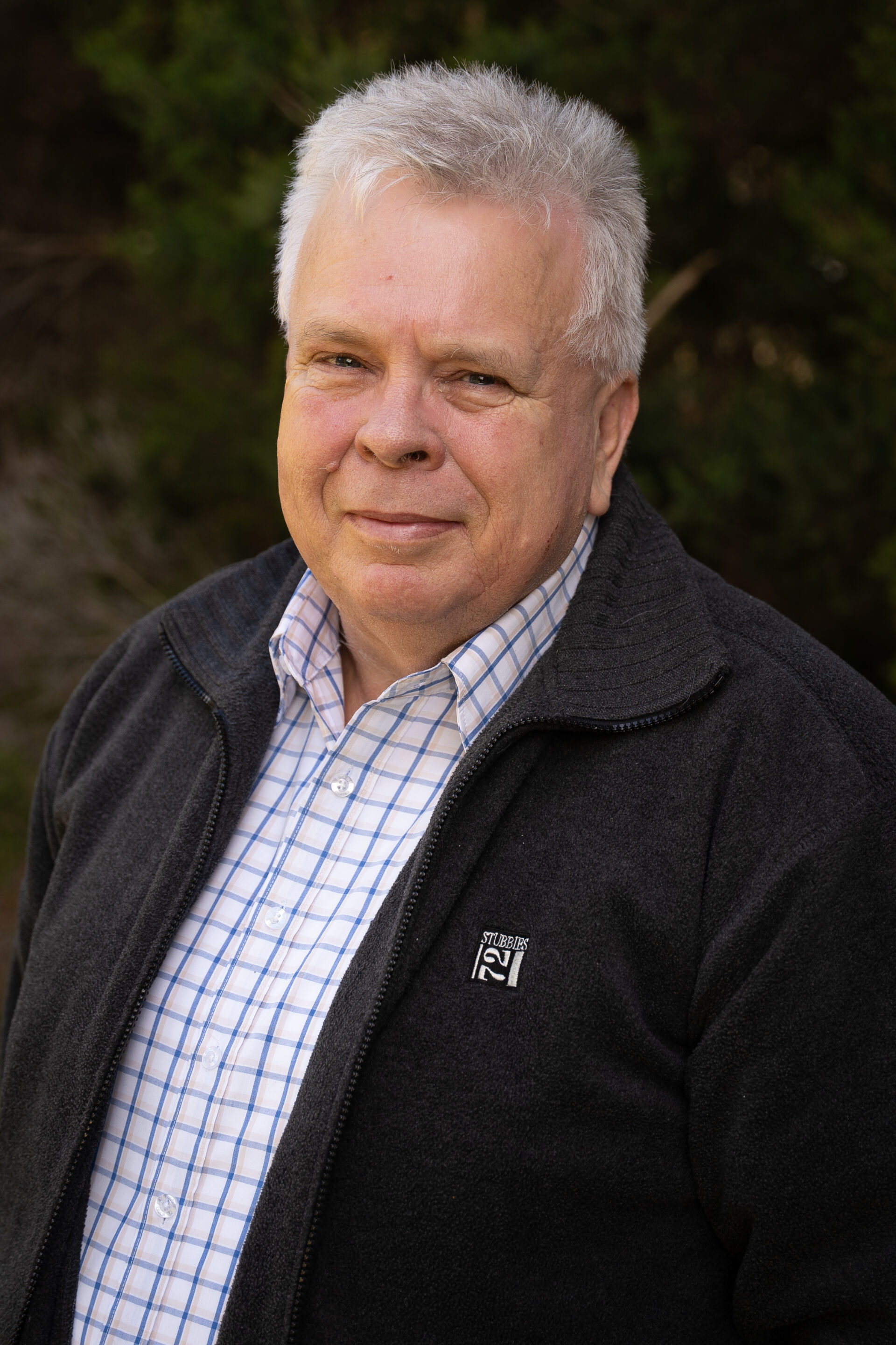 Older man with short grey hair wears a checked shirt and dark jacket, standing outdoors and smiling.