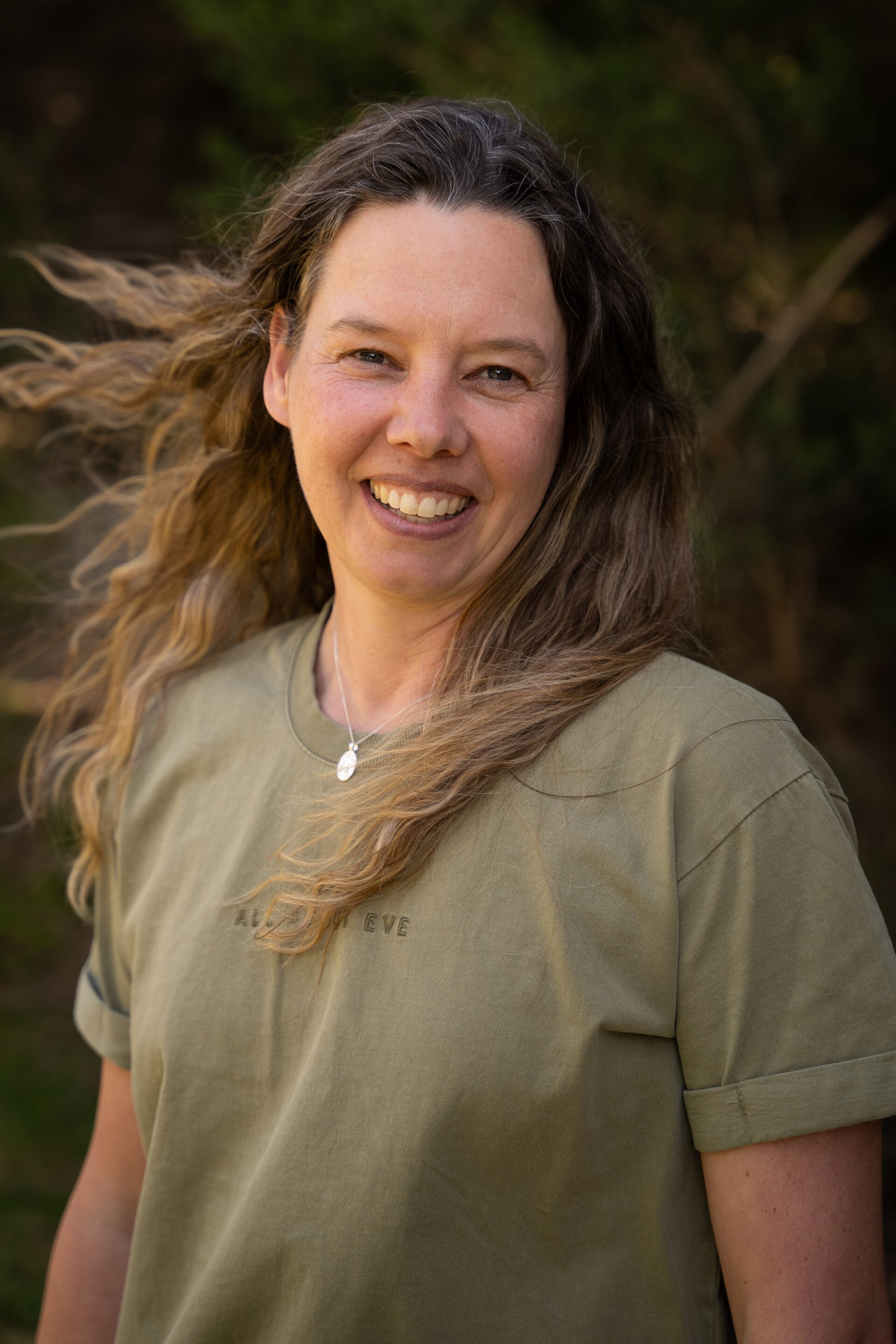 Woman with long wavy hair smiling outdoors, wearing a green t-shirt and necklace.