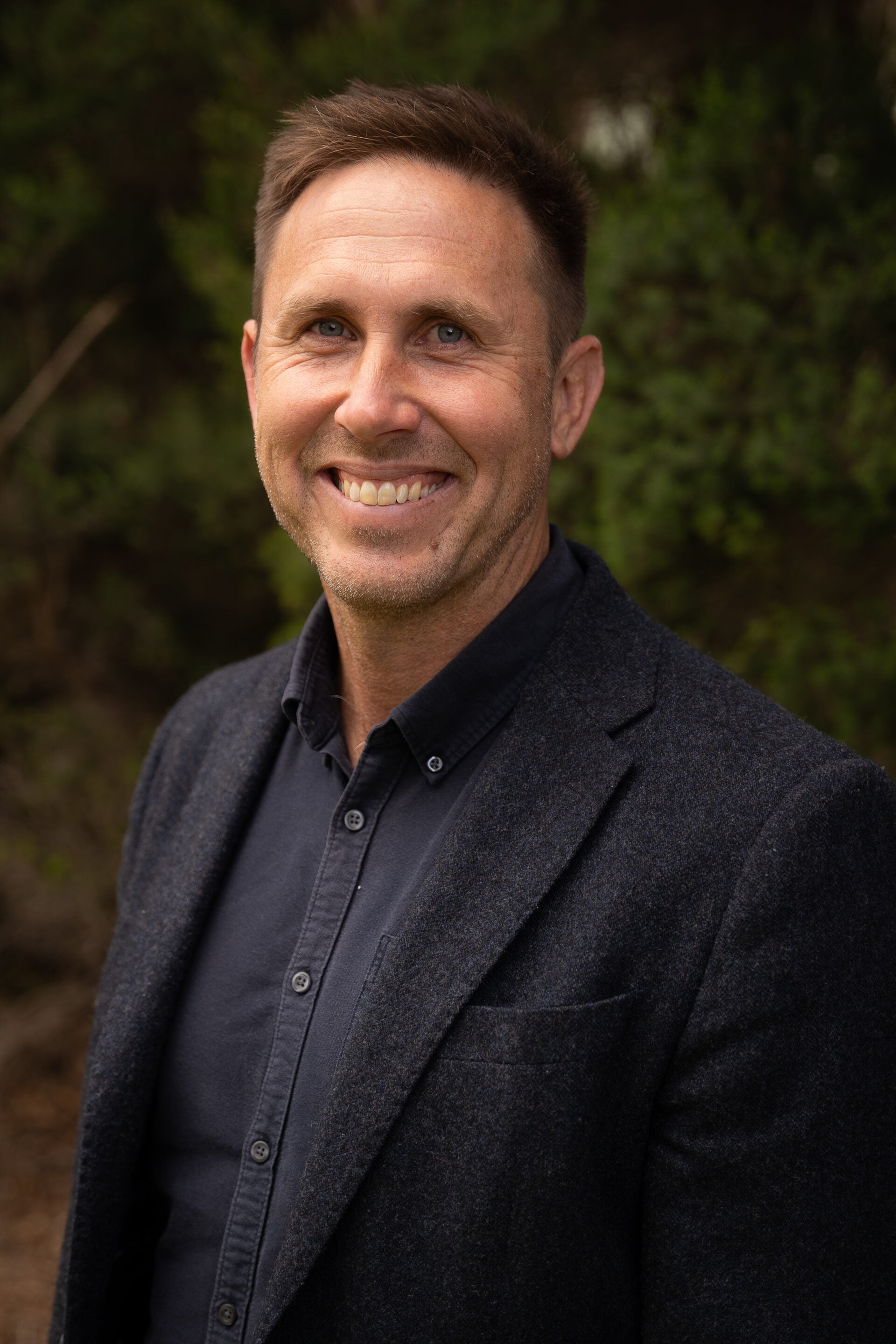 Man in a dark blazer and shirt standing outdoors with greenery in the background.