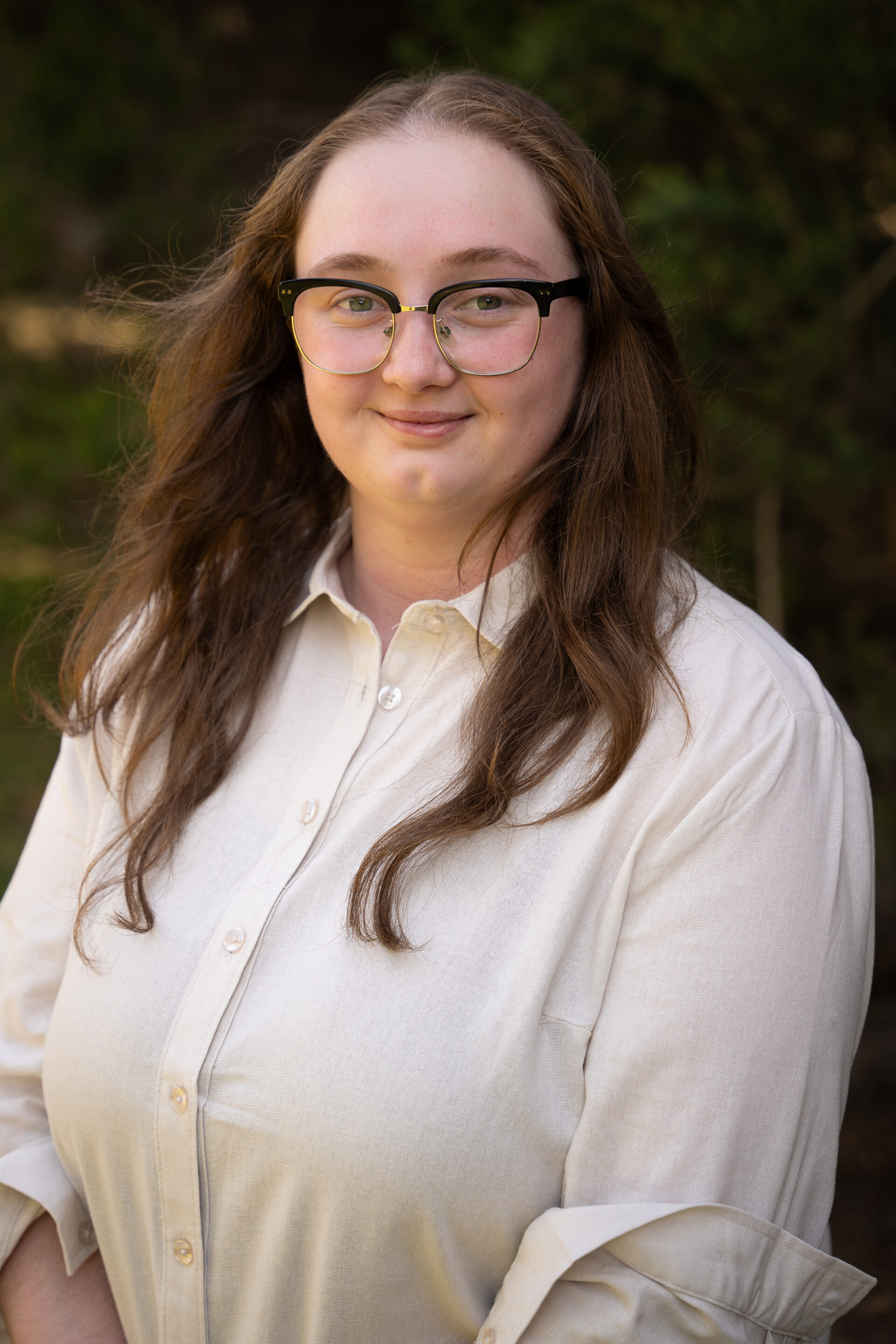 Person with long brown hair and glasses smiling outdoors, wearing a cream-coloured button-up shirt.