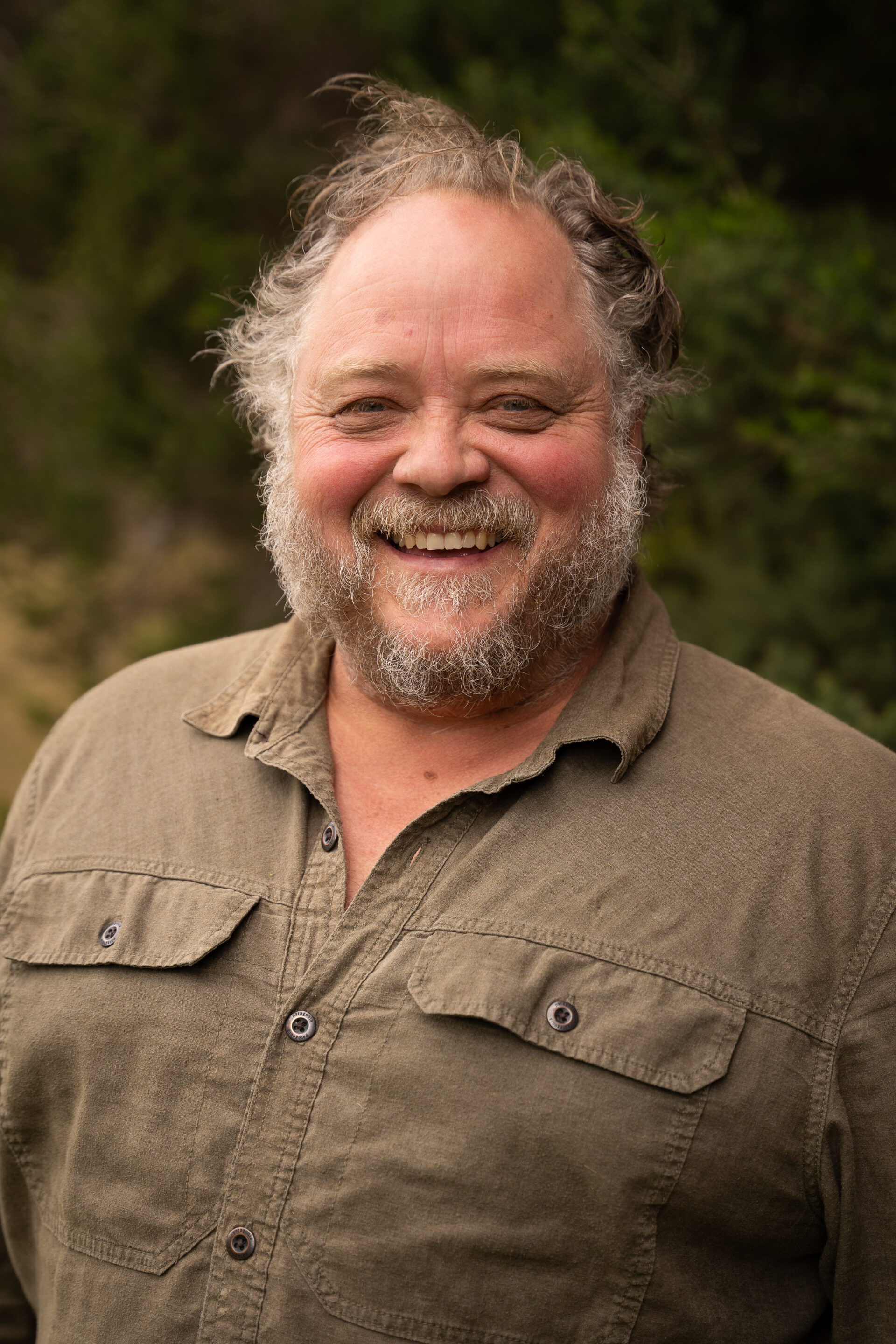 Man with grey beard and khaki shirt standing outdoors in front of green foliage.