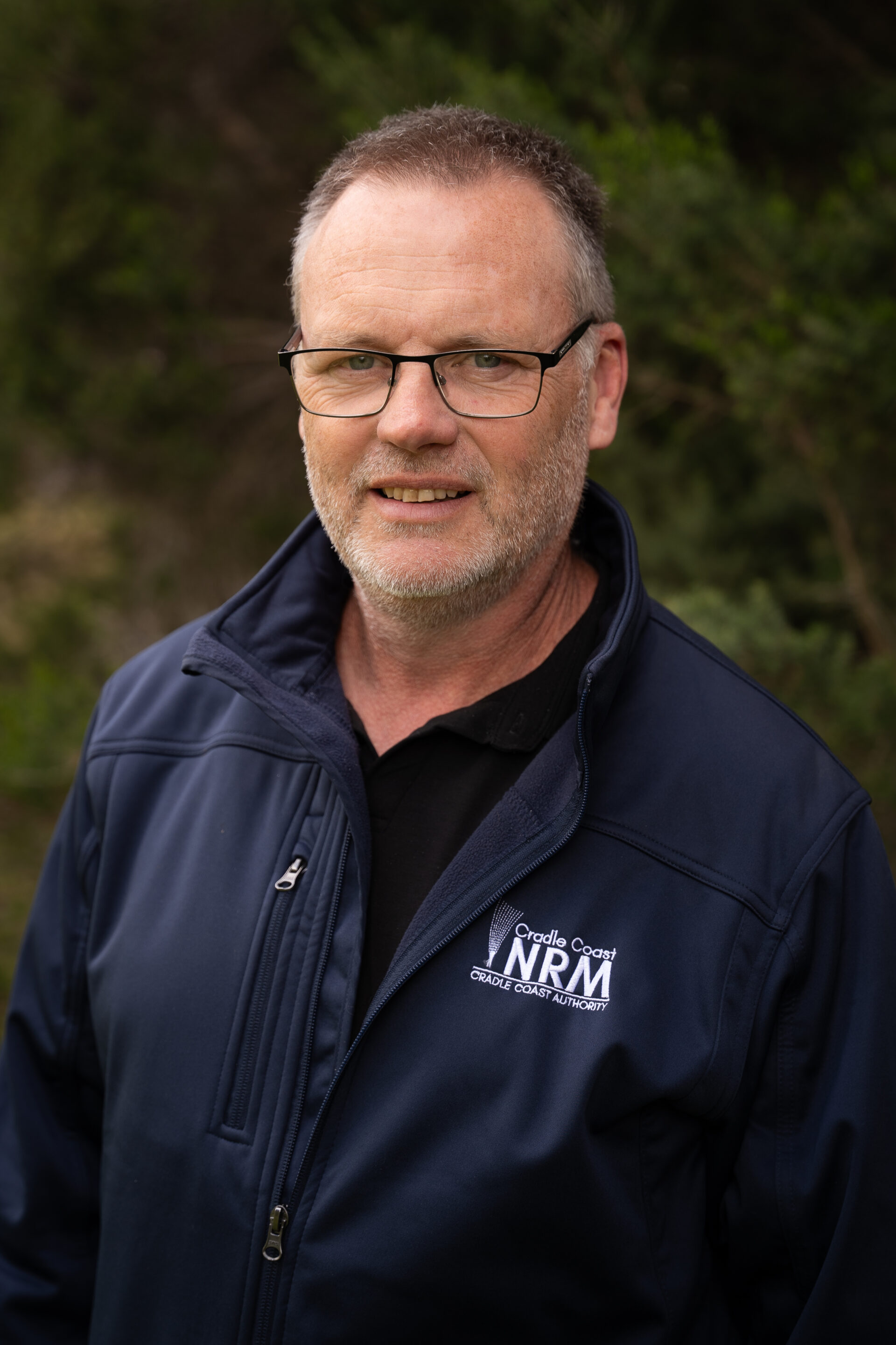 Man with glasses and beard wearing navy jacket standing outside in front of green foliage.