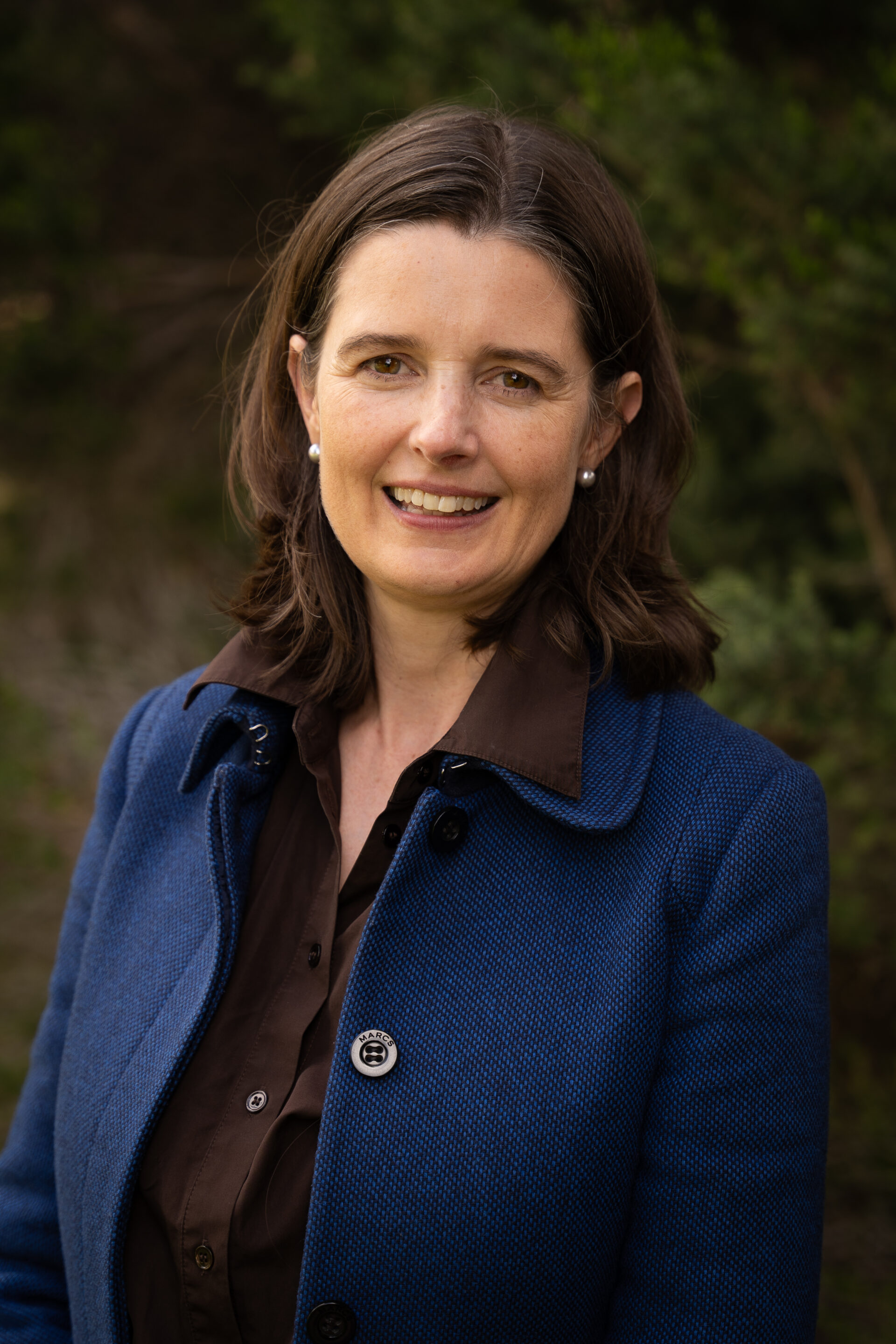 Woman with shoulder-length brown hair, blue coat, and dark shirt smiling outside with greenery in the background.