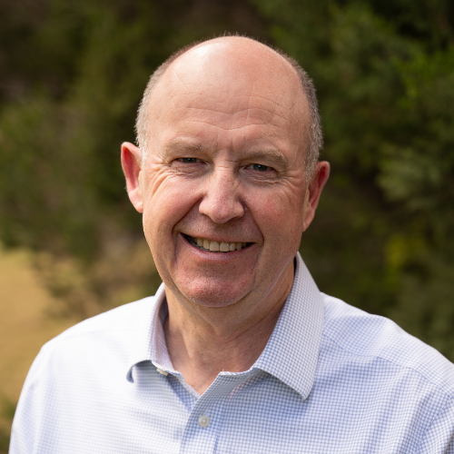 Bald man in a light collared shirt standing outdoors with greenery in the background.