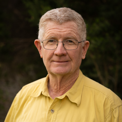 Older man with glasses and short grey hair, wearing a yellow shirt, standing outdoors with greenery in the background.