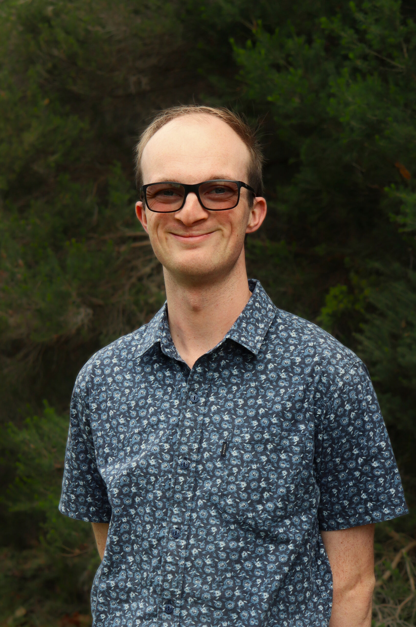 Man with glasses and patterned shirt smiling outdoors in front of green foliage.