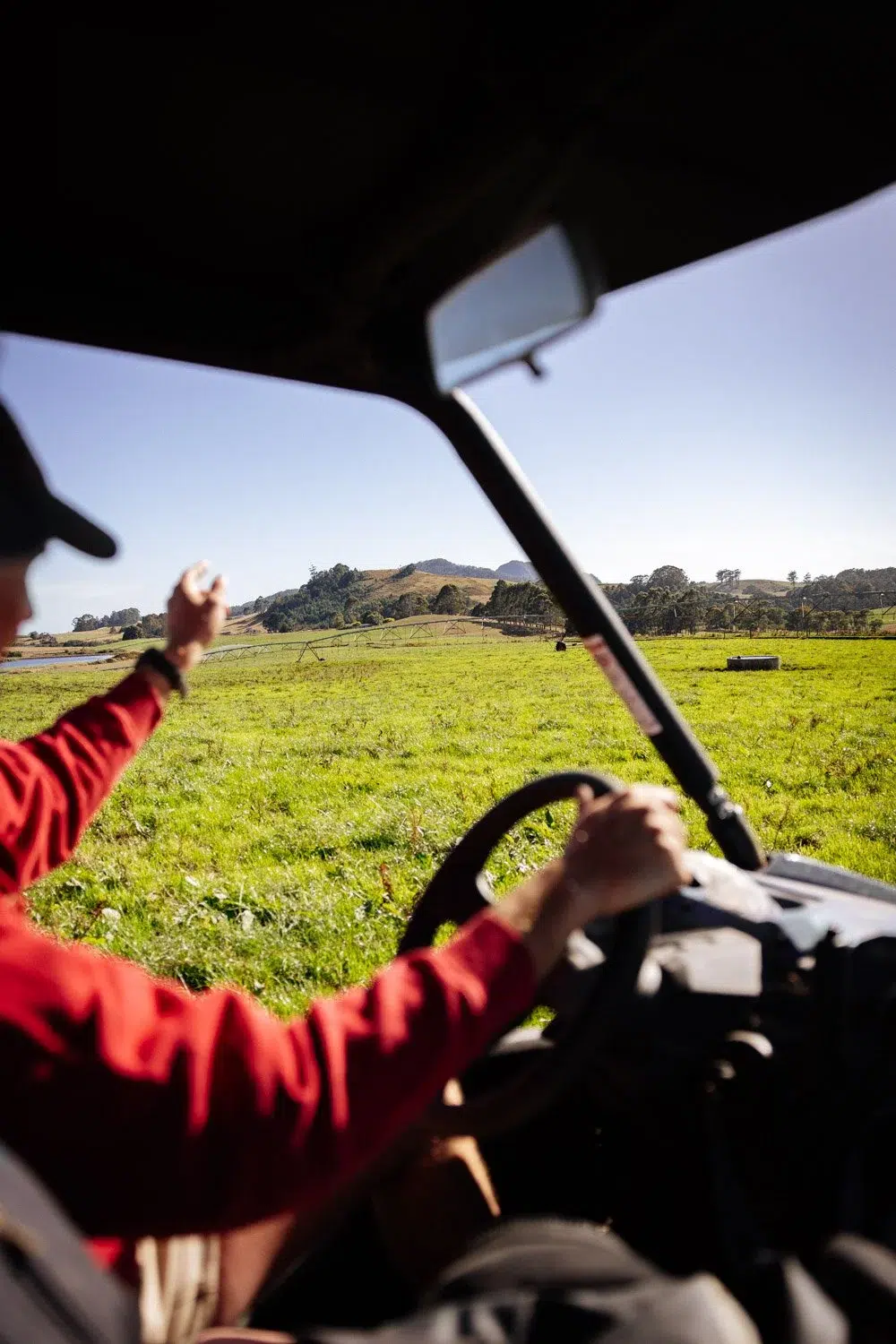 Person driving a vehicle across a green field, waving with one hand on a sunny day.