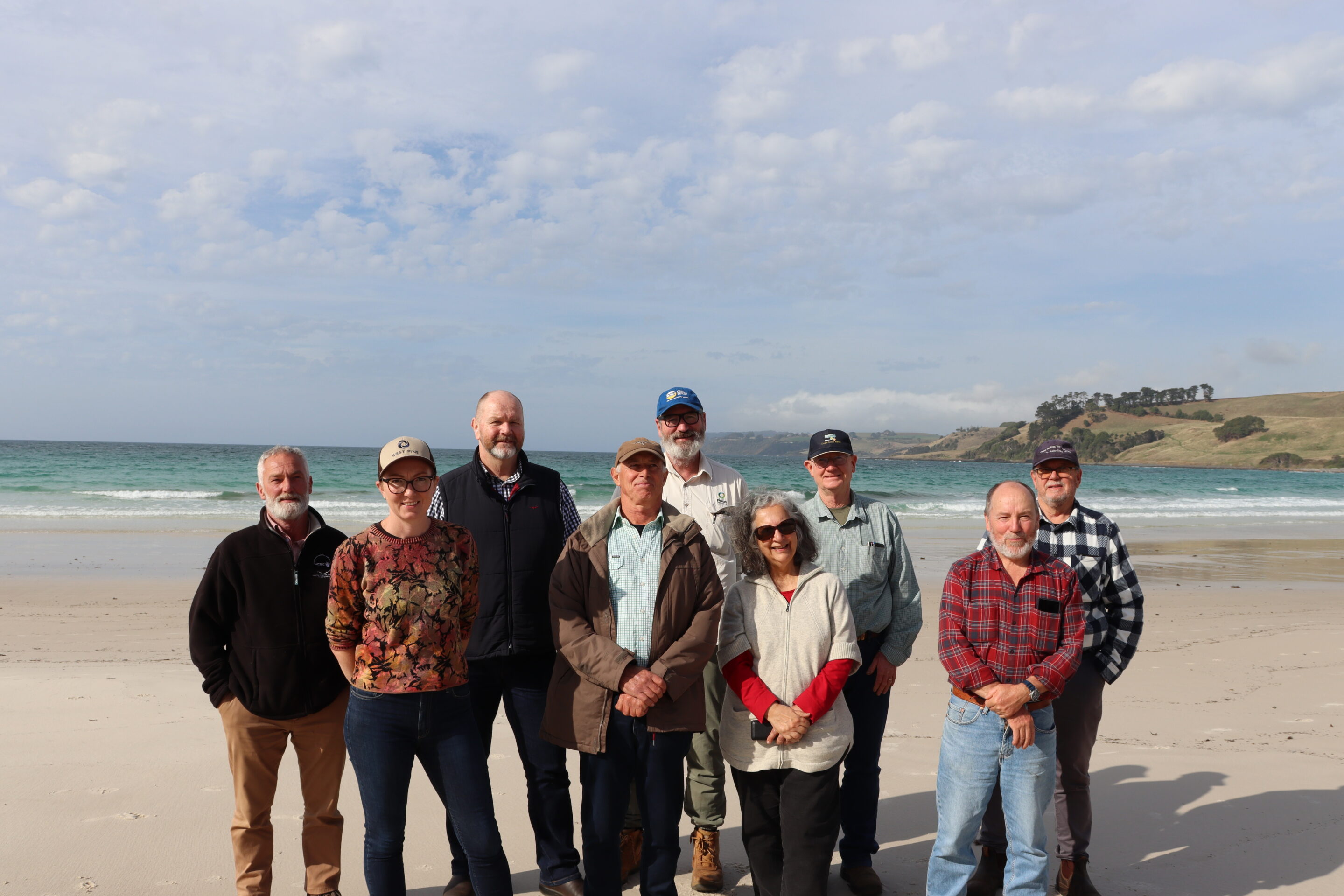 Nine people on a sandy beach with sea waves and hills in the background beneath a partly cloudy sky.