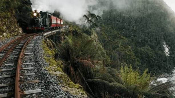 A steam train with red carriages travels on a curved mountain track through dense forest and mist.