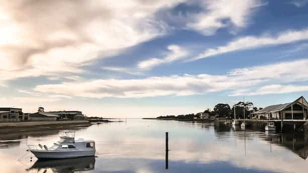 A small white boat on calm water near the shore, with buildings and trees on both sides under a partly cloudy sky.