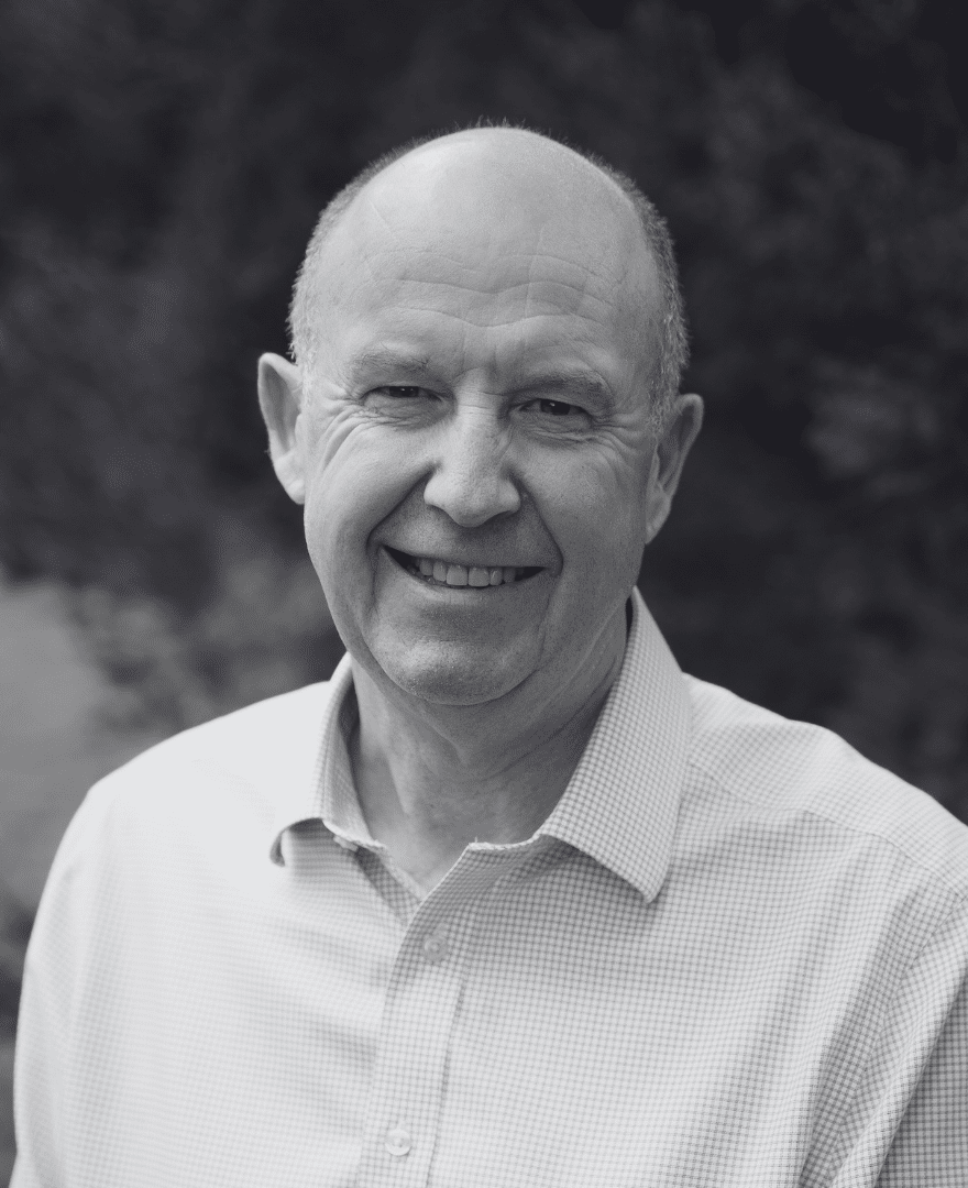 Bald man wearing a light-coloured collared shirt, smiling outdoors with blurred green foliage in the background.